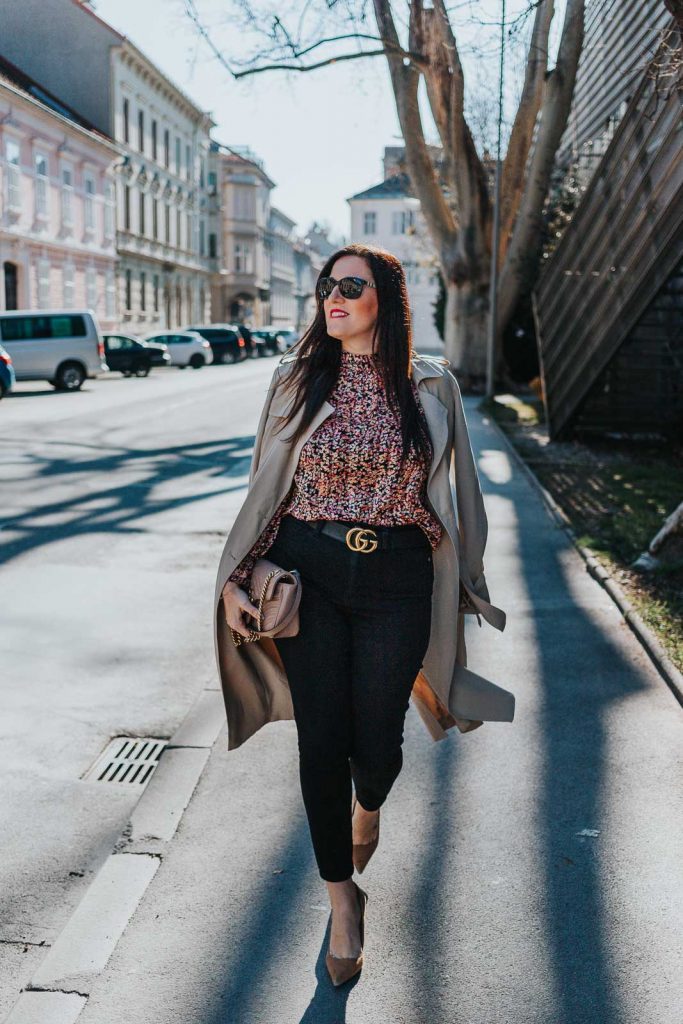 Modebloggerin Miss Classy trägt ein Outfit mit Trenchcoat von Guess, einer schwarzen Skinny Jeans, einer gemusterten Bluse und Pumps. Als Accessoires trägt sie einen Gürtel von Gucci mit Doppel G Schnalle und die Gucci Marmont Schultertasche.