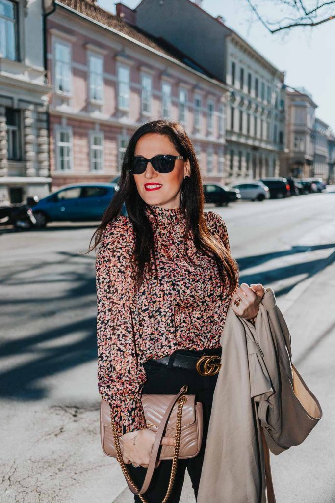 Modebloggerin Miss Classy trägt ein Outfit mit Trenchcoat von Guess, einer schwarzen Skinny Jeans, einer gemusterten Bluse und Pumps. Als Accessoires trägt sie einen Gürtel von Gucci mit Doppel G Schnalle und die Gucci Marmont Schultertasche.