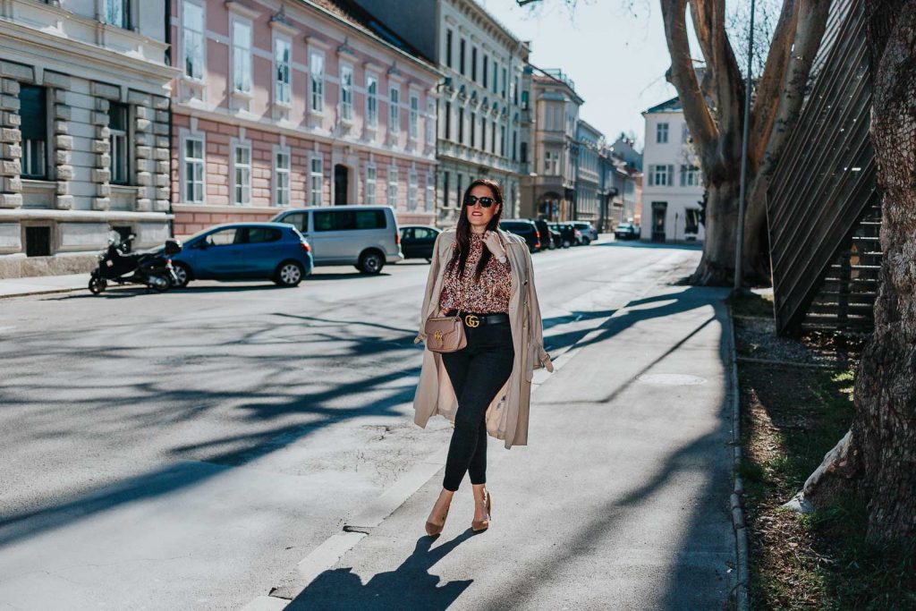 Modebloggerin Miss Classy trägt ein Outfit mit Trenchcoat von Guess, einer schwarzen Skinny Jeans, einer gemusterten Bluse und Pumps. Als Accessoires trägt sie einen Gürtel von Gucci mit Doppel G Schnalle und die Gucci Marmont Schultertasche.