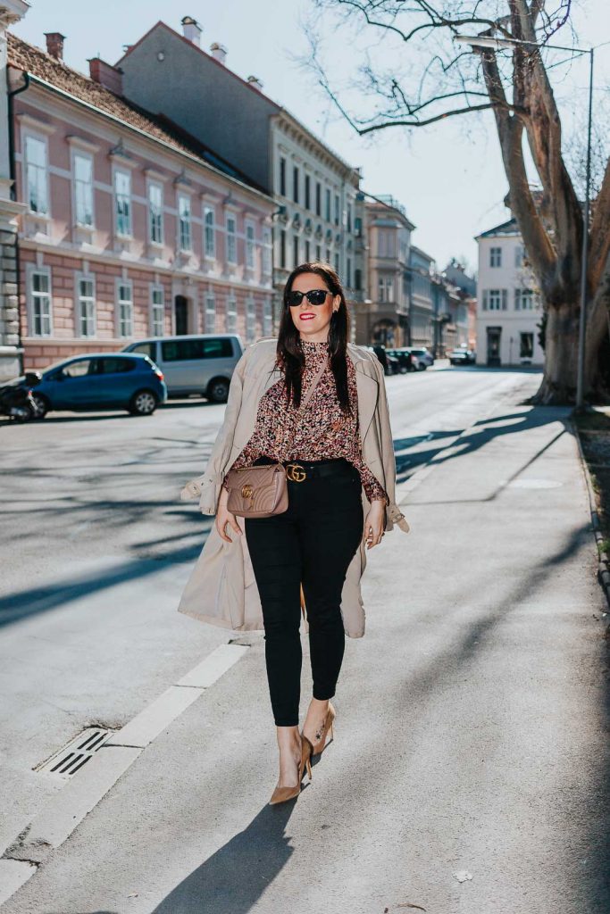 Modebloggerin Miss Classy trägt ein Outfit mit Trenchcoat von Guess, einer schwarzen Skinny Jeans, einer gemusterten Bluse und Pumps. Als Accessoires trägt sie einen Gürtel von Gucci mit Doppel G Schnalle und die Gucci Marmont Schultertasche.