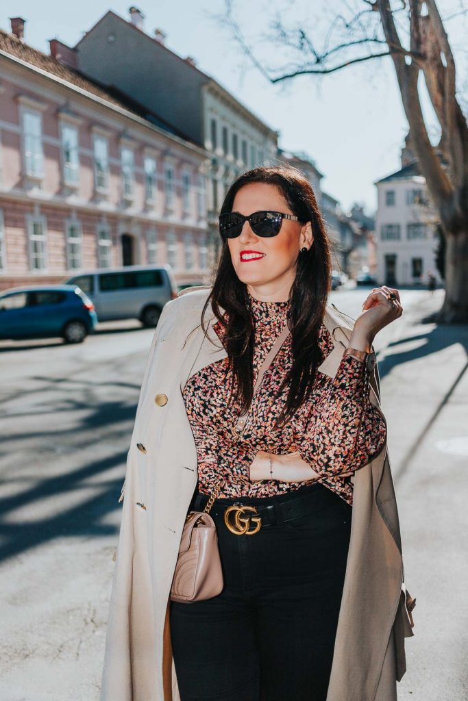 Modebloggerin Miss Classy trägt ein Outfit mit Trenchcoat von Guess, einer schwarzen Skinny Jeans, einer gemusterten Bluse und Pumps. Als Accessoires trägt sie einen Gürtel von Gucci mit Doppel G Schnalle und die Gucci Marmont Schultertasche.