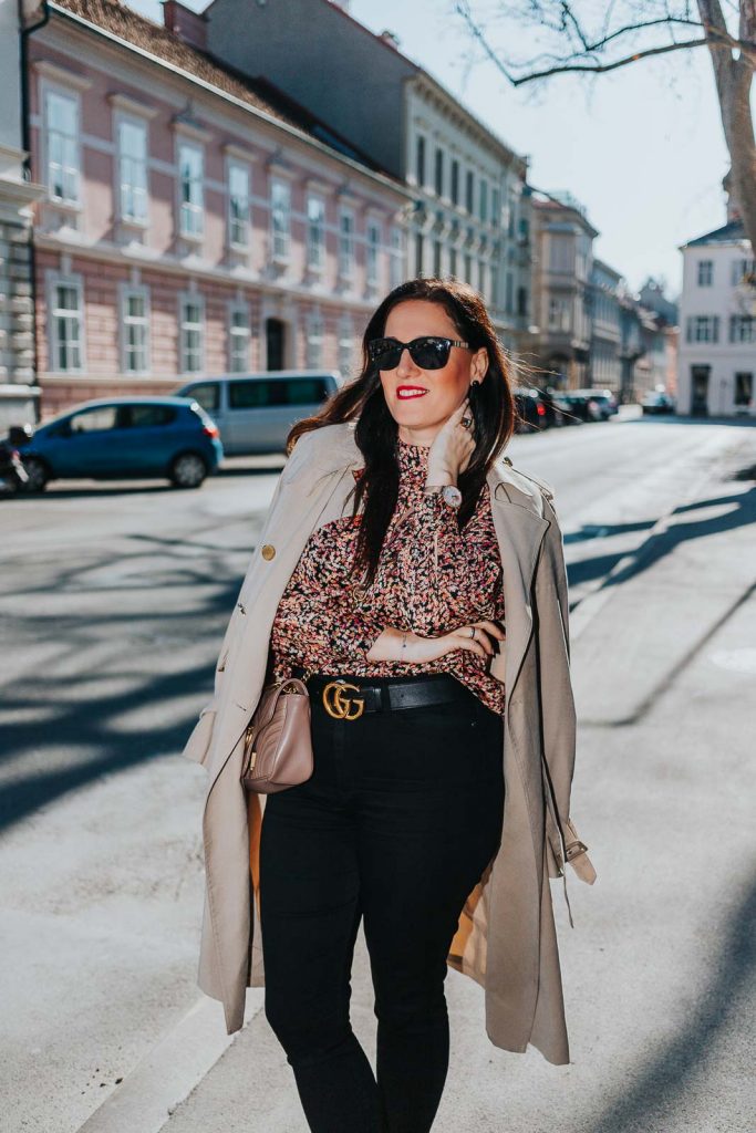 Modebloggerin Miss Classy trägt ein Outfit mit Trenchcoat von Guess, einer schwarzen Skinny Jeans, einer gemusterten Bluse und Pumps. Als Accessoires trägt sie einen Gürtel von Gucci mit Doppel G Schnalle und die Gucci Marmont Schultertasche.